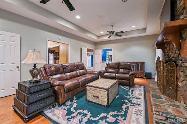 living room with a stone fireplace, hardwood / wood-style floors, ceiling fan, and a tray ceiling