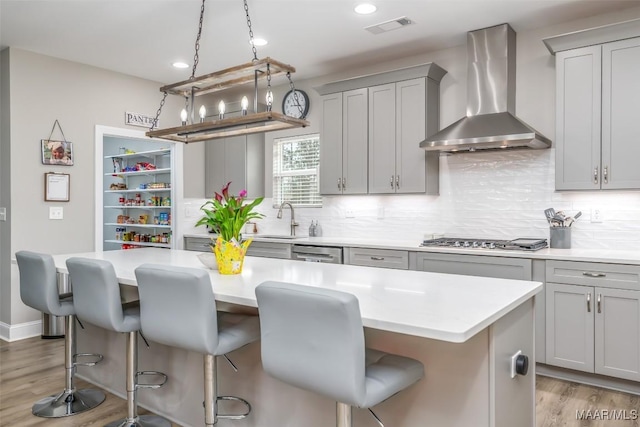 kitchen featuring wall chimney exhaust hood, gray cabinetry, a center island, appliances with stainless steel finishes, and a kitchen breakfast bar