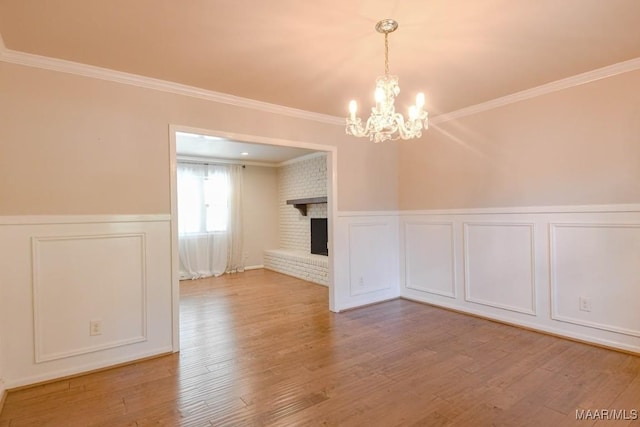 unfurnished dining area with a wainscoted wall, a notable chandelier, ornamental molding, wood finished floors, and a brick fireplace