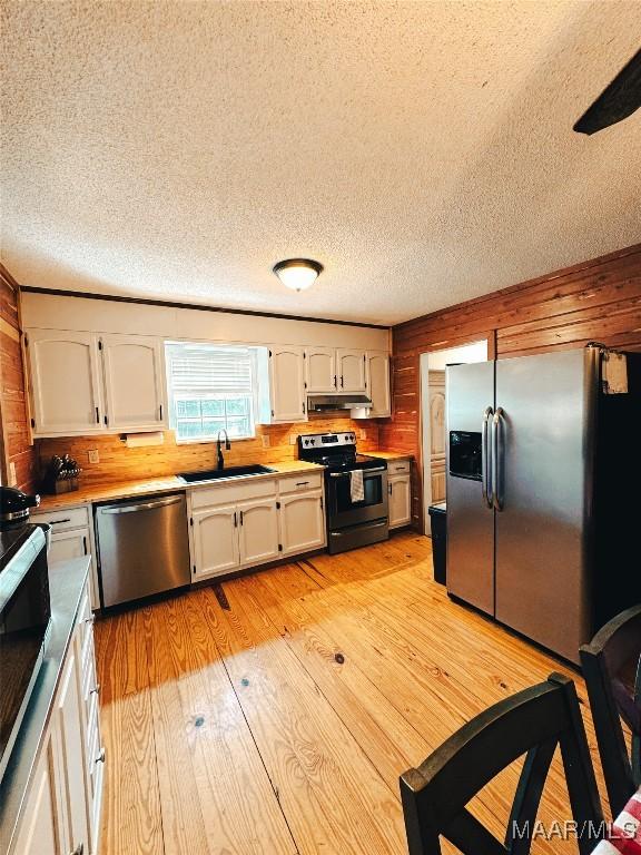 kitchen featuring sink, white cabinetry, light wood-type flooring, appliances with stainless steel finishes, and wooden walls