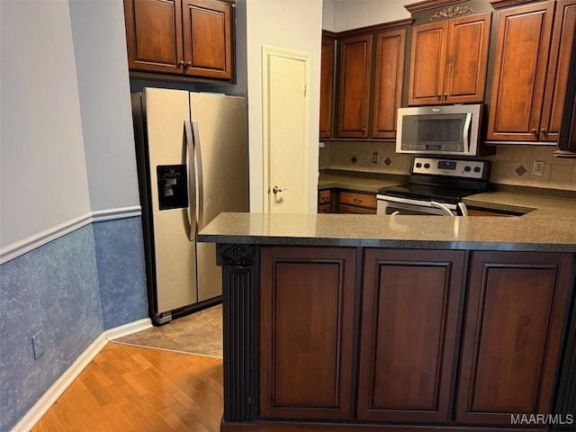 kitchen with dark countertops, a peninsula, stainless steel appliances, light wood-type flooring, and backsplash