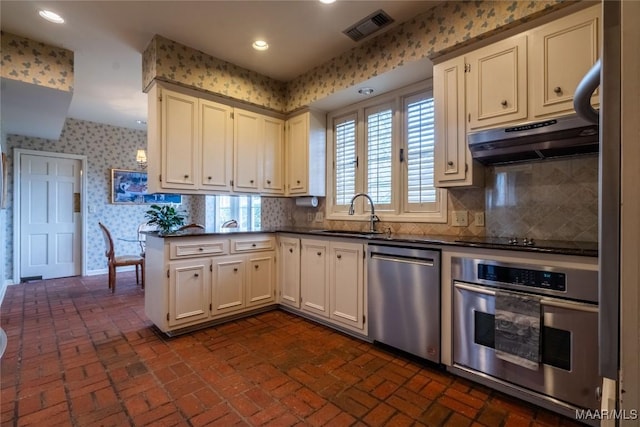 kitchen featuring a sink, dark countertops, under cabinet range hood, appliances with stainless steel finishes, and wallpapered walls