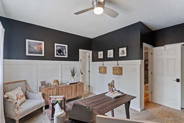 office area with light carpet, ceiling fan, a wainscoted wall, and a textured ceiling