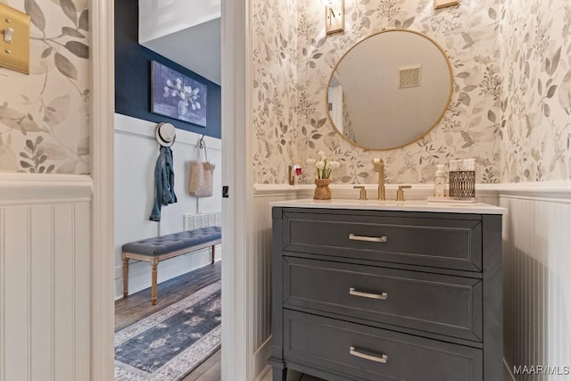 bathroom with a wainscoted wall, visible vents, vanity, wood finished floors, and wallpapered walls