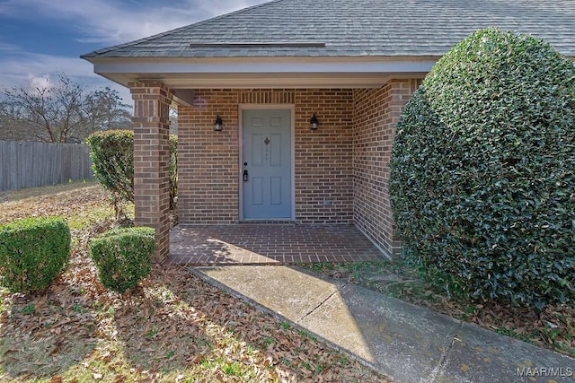 entrance to property featuring a shingled roof, a patio area, brick siding, and fence