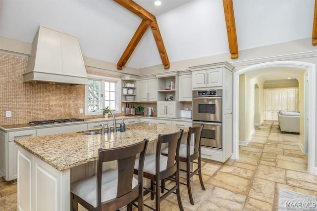 kitchen featuring a center island with sink, stone tile floors, open shelves, custom range hood, and appliances with stainless steel finishes