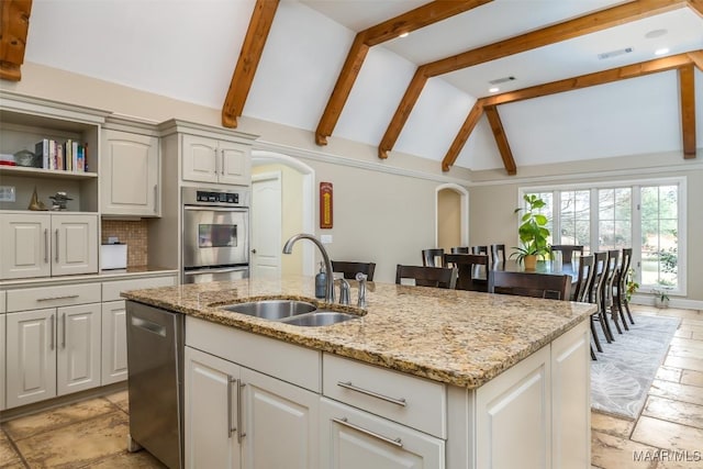 kitchen featuring stone tile floors, a center island with sink, lofted ceiling with beams, appliances with stainless steel finishes, and a sink