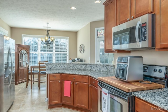 kitchen featuring brown cabinetry, light stone counters, hanging light fixtures, stainless steel appliances, and a textured ceiling