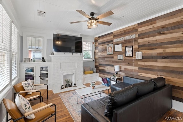 living room featuring ceiling fan, wood walls, wood finished floors, visible vents, and a brick fireplace