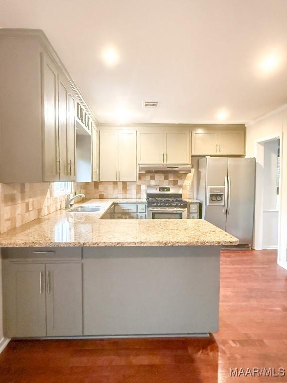 kitchen with appliances with stainless steel finishes, light stone counters, a peninsula, under cabinet range hood, and backsplash