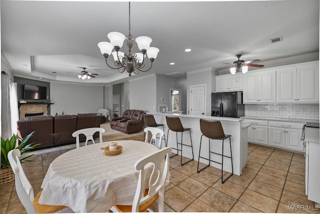 dining space featuring a raised ceiling, light tile patterned flooring, crown molding, a fireplace, and ceiling fan with notable chandelier