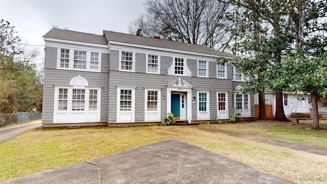 colonial-style house featuring roof with shingles, fence, and a front lawn