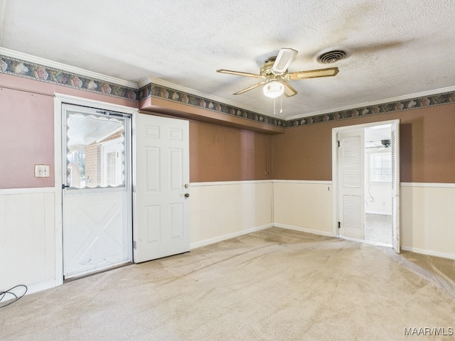 carpeted empty room featuring a textured ceiling, a wainscoted wall, visible vents, a ceiling fan, and crown molding