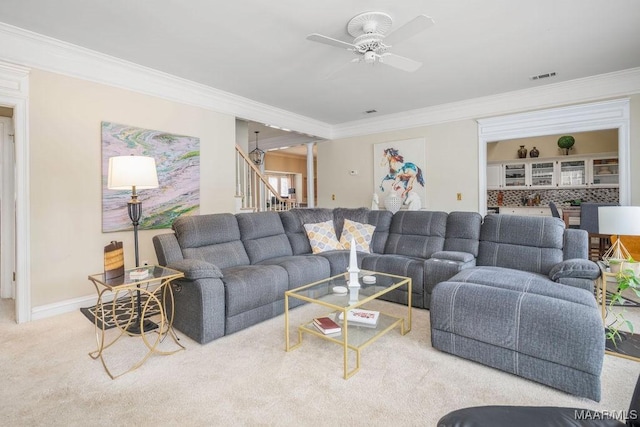 carpeted living room featuring ceiling fan, visible vents, baseboards, ornamental molding, and stairway