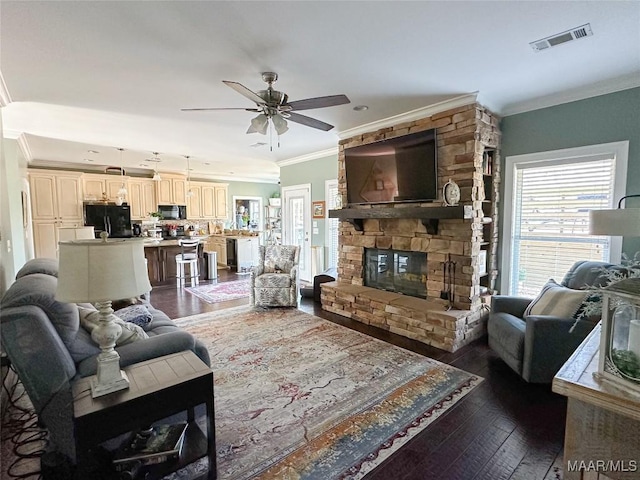 living area featuring visible vents, dark wood finished floors, a ceiling fan, crown molding, and a stone fireplace