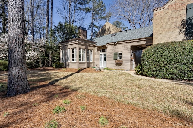 rear view of house featuring french doors, a chimney, a shingled roof, a lawn, and a sunroom