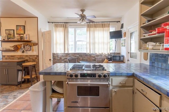 kitchen featuring tile countertops, open shelves, stainless steel appliances, wainscoting, and ceiling fan