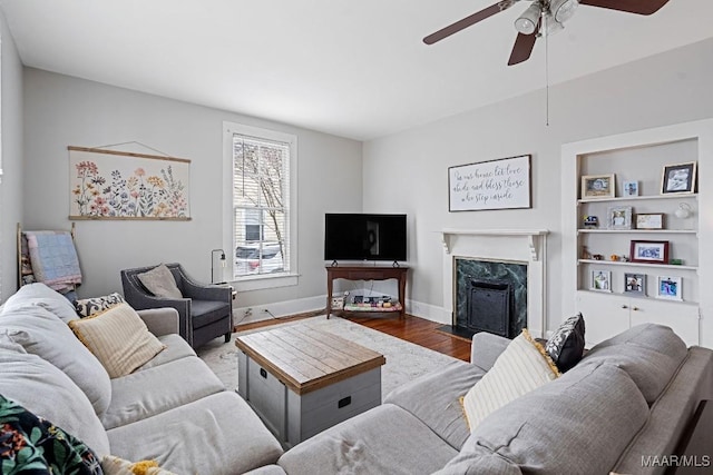 living area with built in shelves, a fireplace, a ceiling fan, wood finished floors, and baseboards