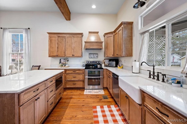 kitchen with beam ceiling, stainless steel appliances, custom range hood, light wood-style flooring, and a sink