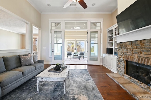 living area featuring baseboards, dark wood-style floors, ceiling fan, crown molding, and a stone fireplace