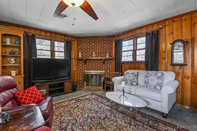 carpeted living area featuring a brick fireplace, visible vents, ceiling fan, and wooden walls