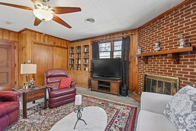 living room featuring wooden walls, carpet flooring, visible vents, ornamental molding, and a brick fireplace