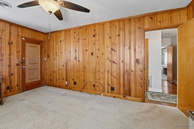 carpeted empty room featuring wood walls, ceiling fan, visible vents, and crown molding