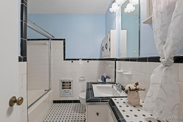 bathroom with toilet, vanity, tile walls, combined bath / shower with glass door, and heating unit