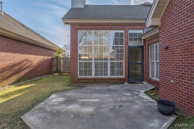 property entrance featuring a shingled roof, brick siding, fence, a yard, and a patio area