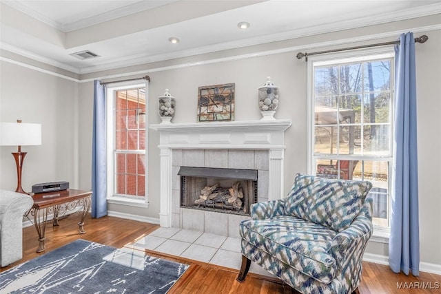 living area with a tiled fireplace, wood finished floors, visible vents, and crown molding