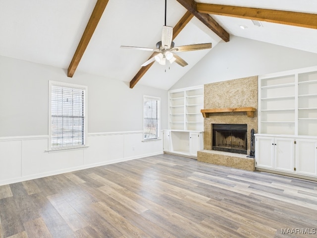 unfurnished living room featuring a ceiling fan, a wainscoted wall, light wood-style flooring, vaulted ceiling with beams, and a fireplace