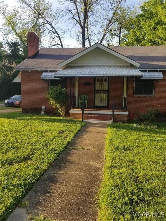 view of front of house featuring covered porch, a chimney, brick siding, and a front lawn