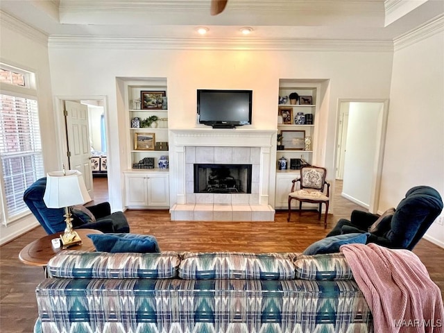 living area featuring crown molding, built in shelves, wood finished floors, a tiled fireplace, and a tray ceiling
