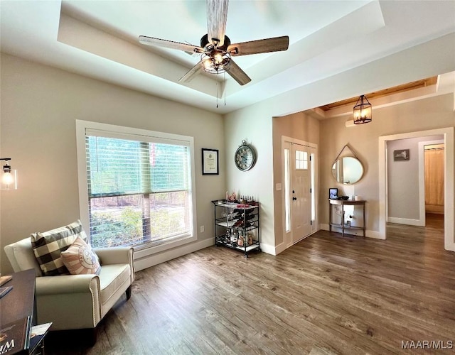 living area featuring a ceiling fan, a raised ceiling, baseboards, and wood finished floors