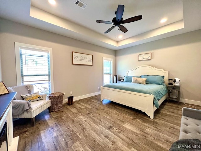 bedroom featuring wood finished floors, visible vents, baseboards, a tray ceiling, and ceiling fan