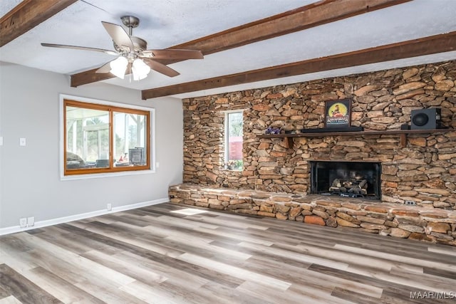 unfurnished living room featuring beamed ceiling, wood finished floors, a stone fireplace, baseboards, and ceiling fan