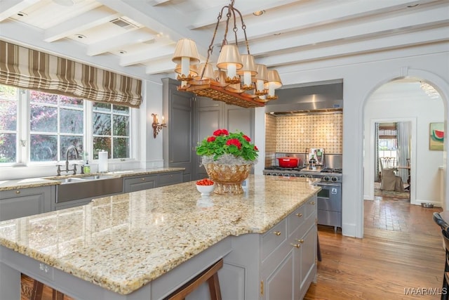 kitchen with beam ceiling, arched walkways, gray cabinets, a sink, and wall chimney range hood