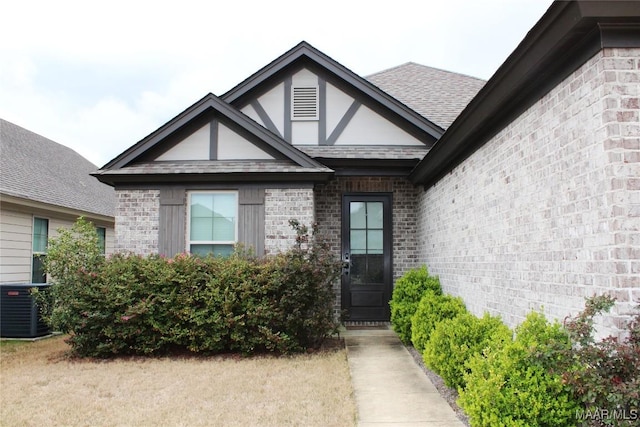 view of exterior entry with central AC unit, brick siding, and roof with shingles