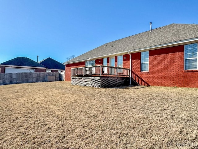 rear view of property featuring fence, roof with shingles, a wooden deck, a lawn, and brick siding