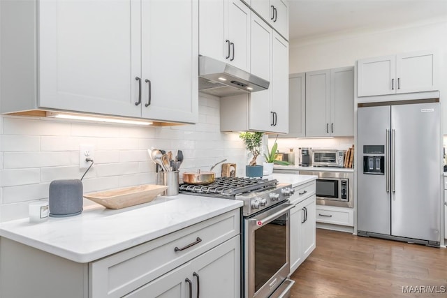 kitchen featuring light stone countertops, wood finished floors, stainless steel appliances, under cabinet range hood, and tasteful backsplash