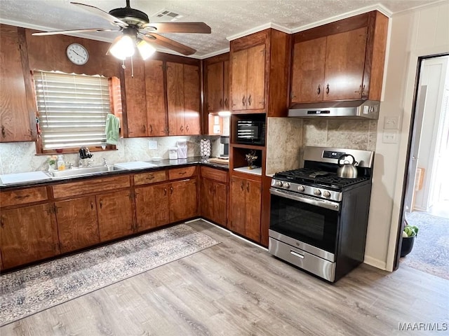 kitchen with a ceiling fan, a sink, under cabinet range hood, stainless steel gas range oven, and brown cabinets