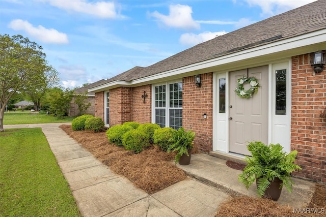 doorway to property featuring a yard, brick siding, and roof with shingles