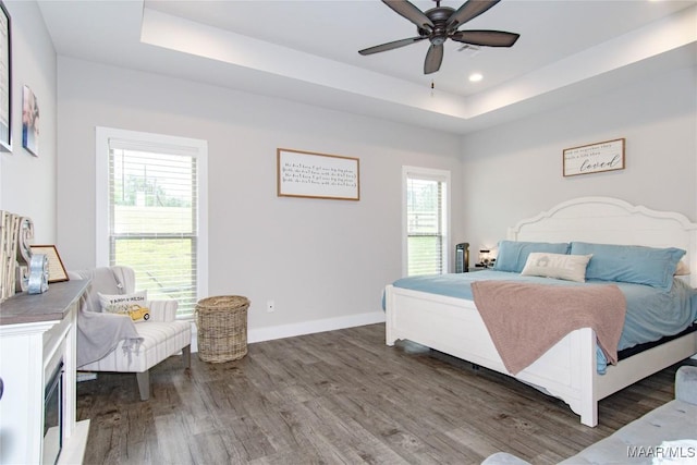 bedroom featuring a tray ceiling, wood finished floors, multiple windows, baseboards, and recessed lighting