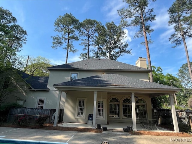 rear view of house featuring a chimney, a shingled roof, covered porch, fence, and stucco siding