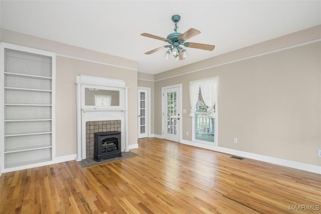 unfurnished living room featuring baseboards, light wood-type flooring, built in features, a wood stove, and ceiling fan