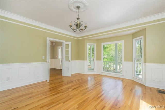 empty room featuring an inviting chandelier, light wood-style flooring, crown molding, a wainscoted wall, and visible vents