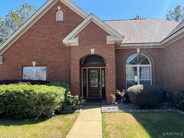 view of exterior entry with roof with shingles, a lawn, and brick siding