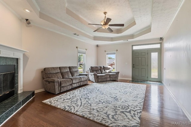 living room with a ceiling fan, a raised ceiling, a tile fireplace, crown molding, and wood finished floors