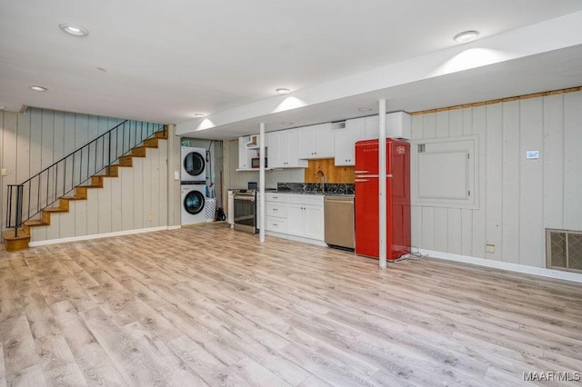 unfurnished living room featuring stacked washer and clothes dryer, light wood-type flooring, baseboards, stairs, and wooden walls