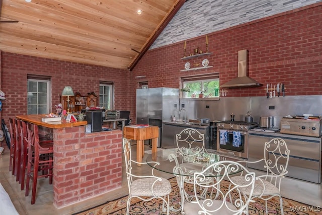 kitchen with wooden ceiling, appliances with stainless steel finishes, wall chimney range hood, a sink, and high vaulted ceiling
